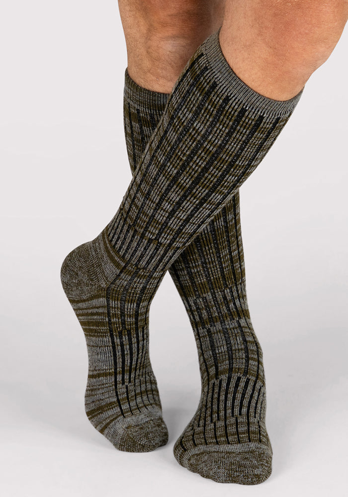 A pair of legs wears Woolx Mens Joey Mid Calf Socks in Dark Forest Marled, featuring ribbed texture and a gray and olive green plaid pattern, shown against a plain light background.
