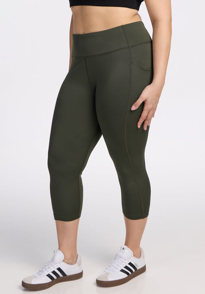 A person wearing Woolx Merino Flex® McKenna Capris in Everleaf and white Adidas sneakers with black stripes stands against a plain white background, showing only the lower torso and legs. | Juliana is 5'10", wearing a size XL