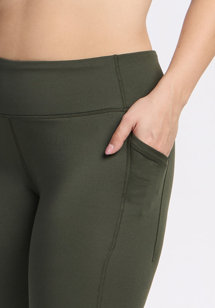 A person wears Woolx Merino Flex® McKenna Capris in Everleaf (olive green) with a hand in the side pocket. The image is cropped to highlight the lower torso and upper legs against a plain background.