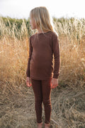 A child standing in a field, wearing a brown thermal top and pants.