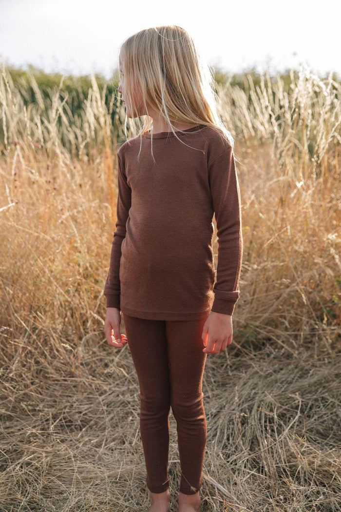 A child standing in a field, wearing a brown thermal top and pants.