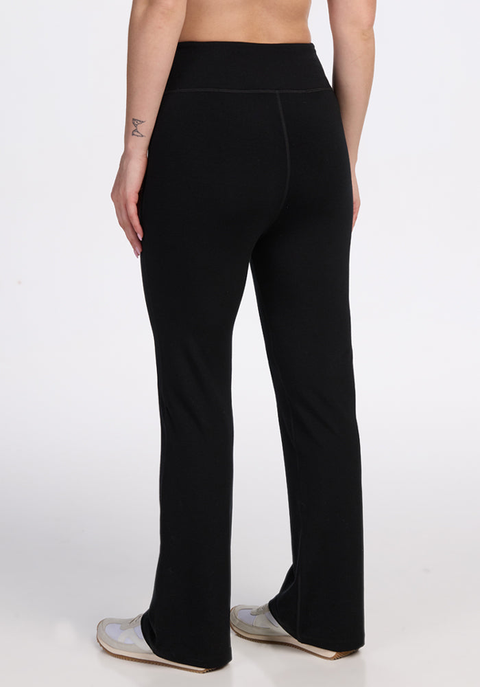 A person stands facing away, wearing Woolx Morgan Flare Pants - Black with a high-waisted, wide-leg fit and white shoes. Only the lower back, legs, and midriff are visible against a plain white background.