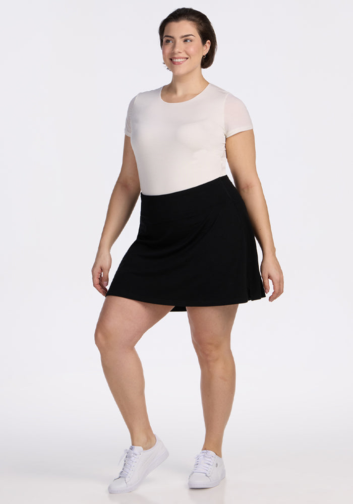 A woman stands smiling against a plain white background, wearing a short-sleeved white top, the Woolx Sienna Skort - Black with pockets, and white sneakers. | Juliana is 5'10", wearing a size XL