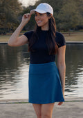 A woman wearing a white cap, black t-shirt, and Woolx Sienna Skort in Majolica Blue smiles by a peaceful lakeside in the park, with trees and grass visible in the background.